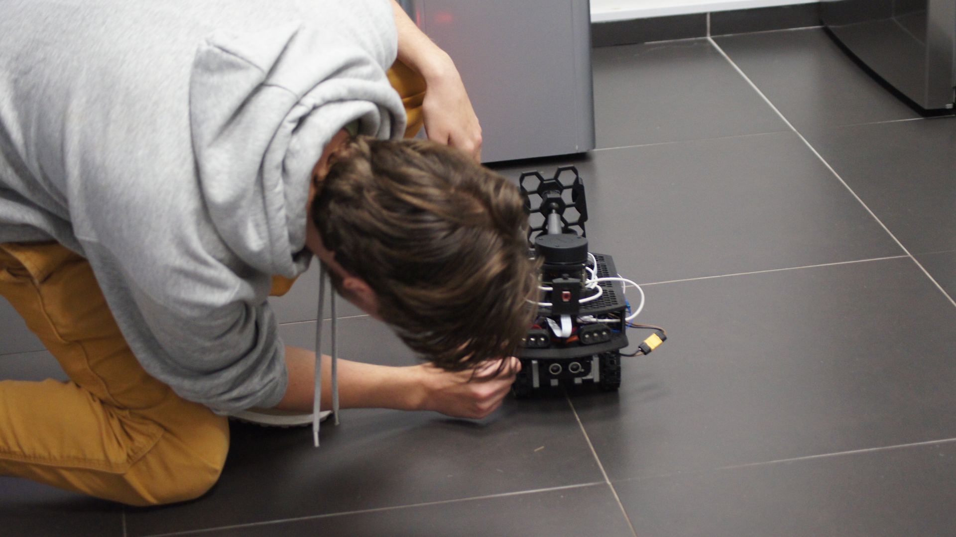 Tracked robot on dark tile floor, white LED headlights active, viewed from above at angle — 3D-printed chassis with hexagonal antenna mount and camera