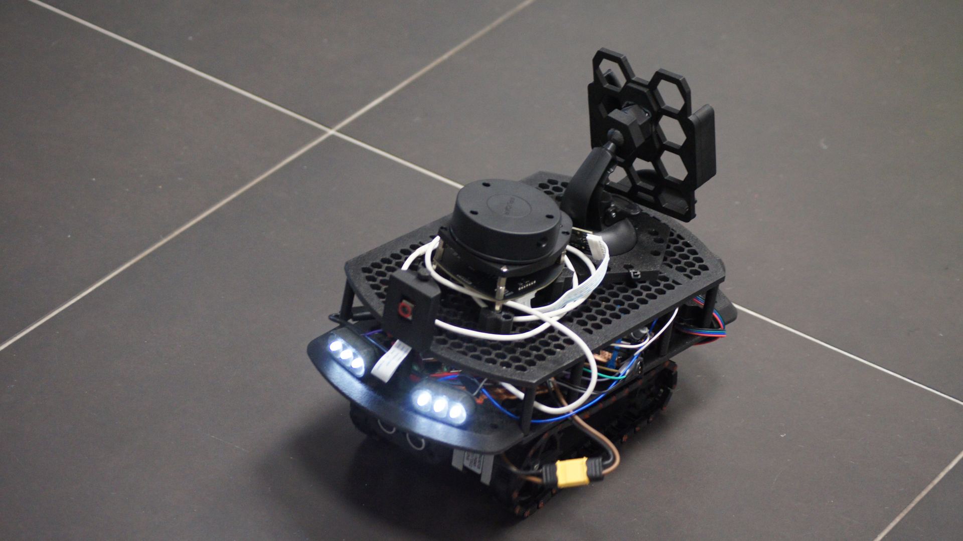 Tracked robot front view on dark tile floor, white LED headlights on, ultrasonic sensors visible on front bumper, XT60 power connector on side